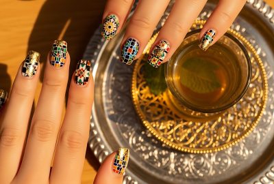 Close-up of elegant hands with unique mosaic nail art holding a glass of traditional Moroccan mint tea, set on a decorative silver and gold tray, showcasing style and cultural richness.