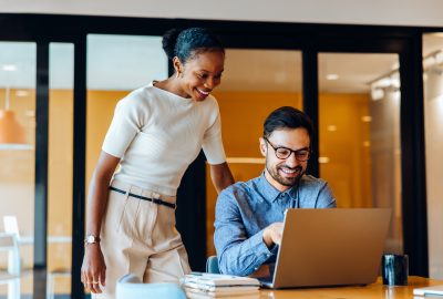A cheerful man and woman work together on a laptop in a professional office setting. They appear to be discussing a shared project, fostering teamwork while enjoying their workplace environment.