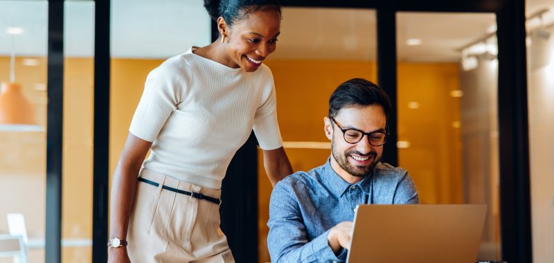 A cheerful man and woman work together on a laptop in a professional office setting. They appear to be discussing a shared project, fostering teamwork while enjoying their workplace environment.