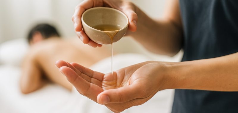 Horizontal intimate spa moment with warm massage oil being poured into the therapist’s hands as a client rests in the blurred background captured in natural soft lighting to express warmth, relaxation
