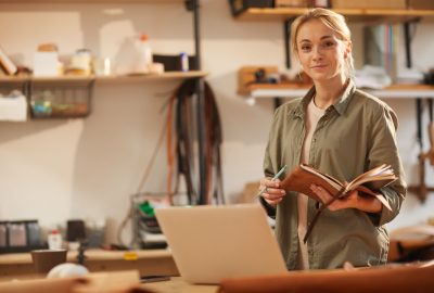 Horizontal portrait of attractive young woman wearing casual outfit standing at table with laptop on it holding notebooks looking at camera