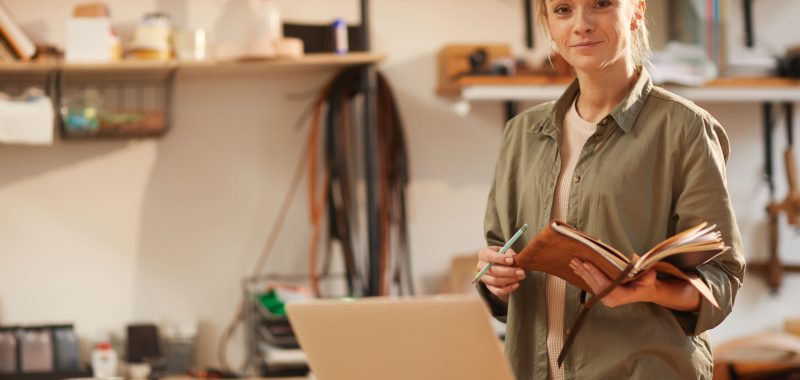 Horizontal portrait of attractive young woman wearing casual outfit standing at table with laptop on it holding notebooks looking at camera