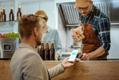 Food Truck Employee Hands Out a Freshly Made Burger to a Happy Young Male. Man in a Casual Suit is Using His Smartphone for NFC Mobile Payment Solution. Street Food Truck Selling Burgers.