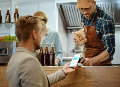 Food Truck Employee Hands Out a Freshly Made Burger to a Happy Young Male. Man in a Casual Suit is Using His Smartphone for NFC Mobile Payment Solution. Street Food Truck Selling Burgers.