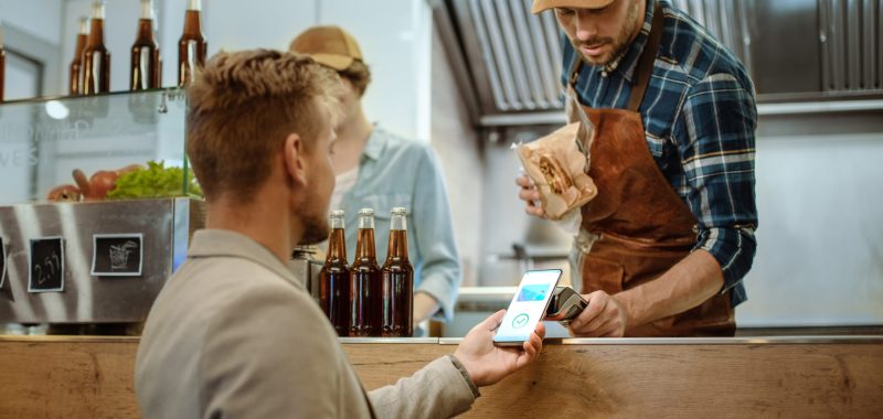 Food Truck Employee Hands Out a Freshly Made Burger to a Happy Young Male. Man in a Casual Suit is Using His Smartphone for NFC Mobile Payment Solution. Street Food Truck Selling Burgers.