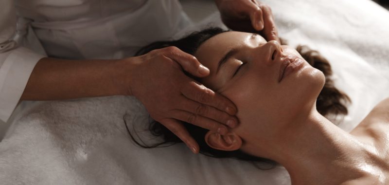 Young woman enjoying massage at spa salon. Beautician rubbing temples of client, smiling girl relaxing during myofascial massaging therapy at beauty clinic, cosmetology procedure.