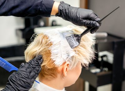 Coloring white hair with hair dye and brush by hands of hairstylist for the young caucasian blonde woman at a hair salon, close up