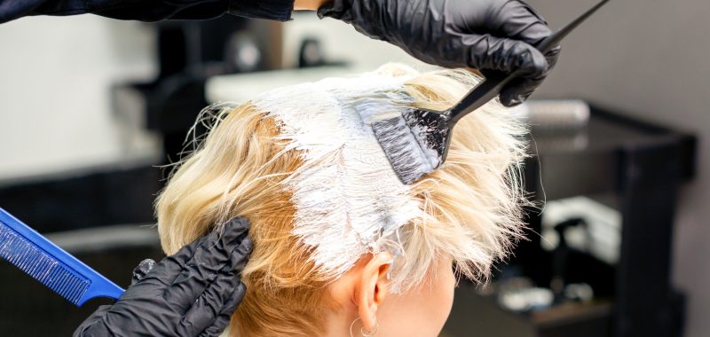 Coloring white hair with hair dye and brush by hands of hairstylist for the young caucasian blonde woman at a hair salon, close up