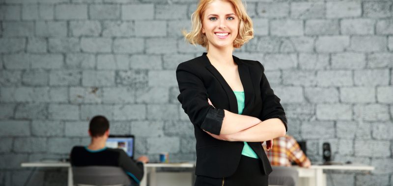 Young happy businesswoman with arms folded standing in office