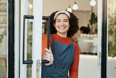 Portrait, welcome and woman at coffee shop door for opening with friendly smile of hospitality. Cafe, entrance and retail with happy restaurant owner or waitress at store for greeting or service.