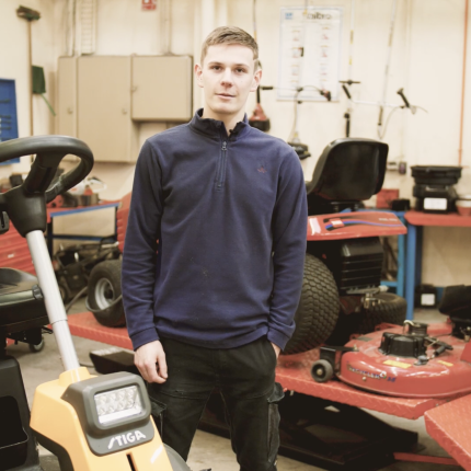 Jeune-homme apprenant la mécanique en matériels d'espace vert posant devant des engins dans un atelier.