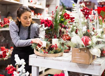 Fleuriste créant une décoration florale de Noël tendance 2025 avec bougies, pommes de pin et poinsettias.