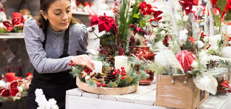 Fleuriste créant une décoration florale de Noël tendance 2025 avec bougies, pommes de pin et poinsettias.
